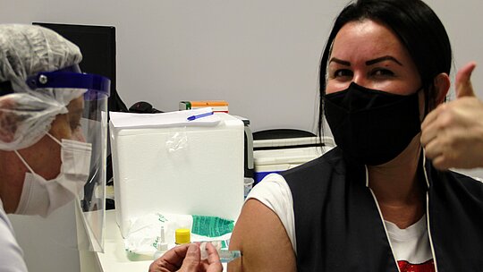 mulher morena, de máscara e com colete e crachá do MAckenzie, recebe injeção de outra mulher paramentada com máscara, jaleco e touca.