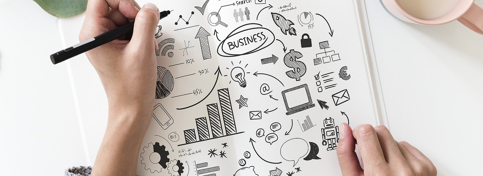 uma mão com um lápis sobre uma mesa. na folha de papel tem muitos desenhos referentes à economia