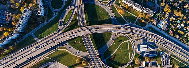 Foto aérea mostra estradas e diversos carros. Tem um pouco de verde e também algumas casas prédios, mas o foco são as vias com veículos.