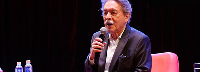 A foto mostra o arquiteto Paulo Mendes da Rocha em um evento do Mackenzie, no ano de 2017. Ele está com um blazer e segurando um microfone, sentado em uma poltrona vermelha. 
