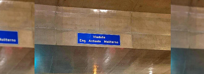Na imagem, é possível ver a lateral de um viaduto na cor cinza concreto com uma placa de fundo azul com letras brancas onde é possível ler "Engenheiro Antonio Moliterno"