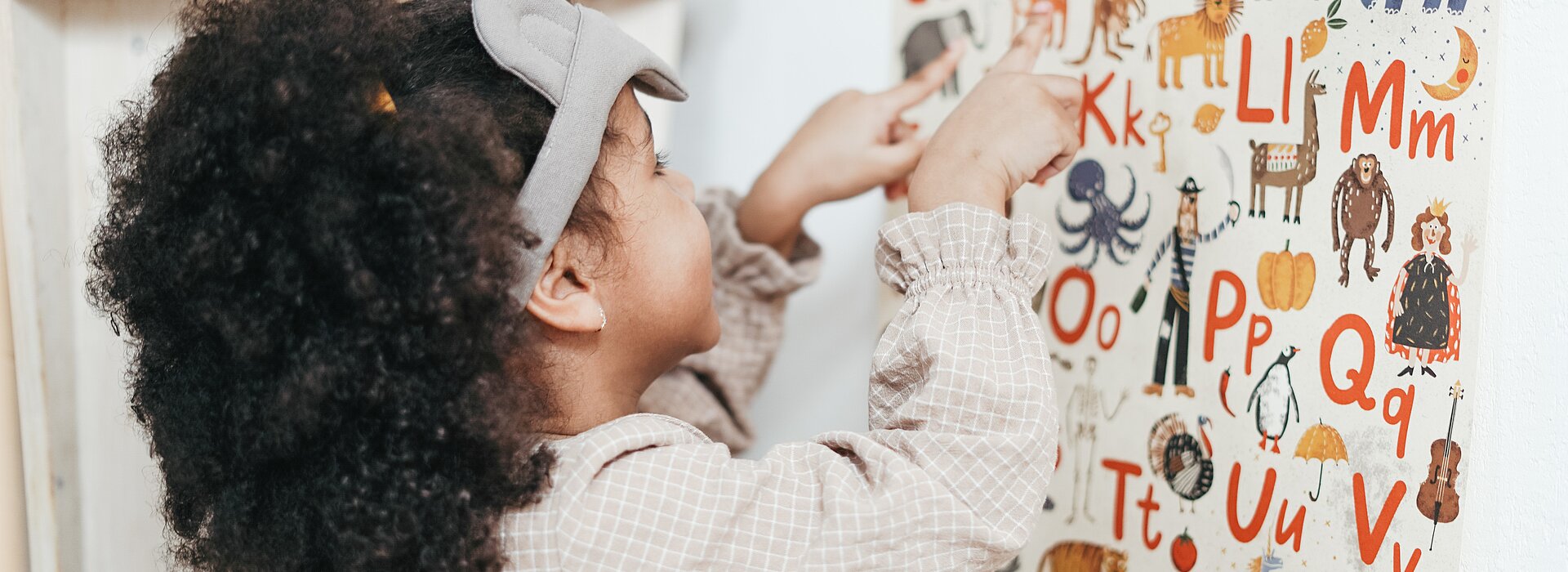 Uma menina de aproximadamente quatro anos está em frente a um painel cheio de letras do alfabeto. Todas as letras têm ao lado um animal, como girafa, elefante. 