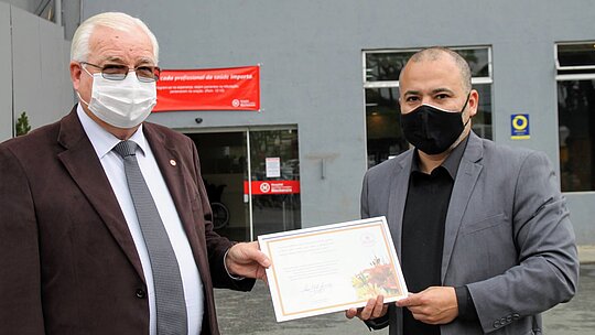 Dois homens com máscaras e ternos seguram um quadro de reconhecimento em frente ao hospital