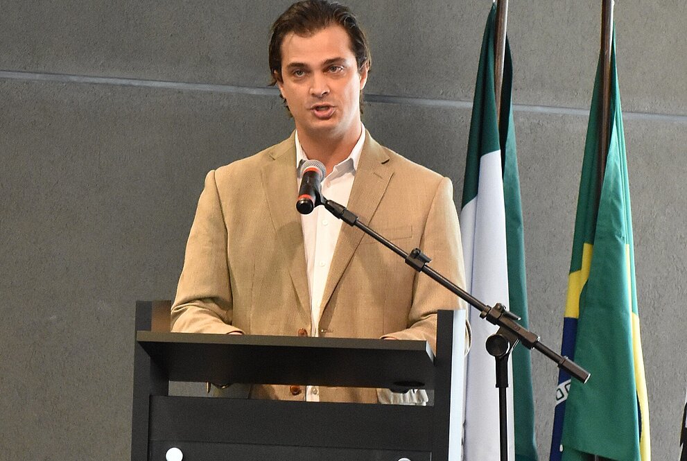 Pedro Buck está falando no pulpito em cima do palco. Atrás dele está a bandeira do Brasil.
