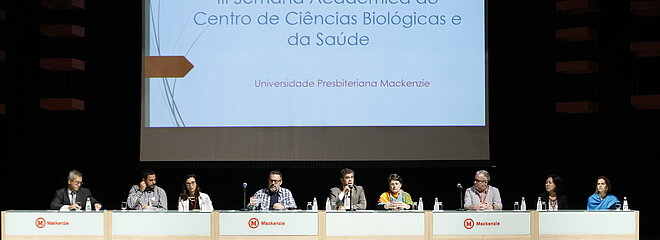 autoridades sentadas à mesa durante evento. Telão na parte de trás da imagem