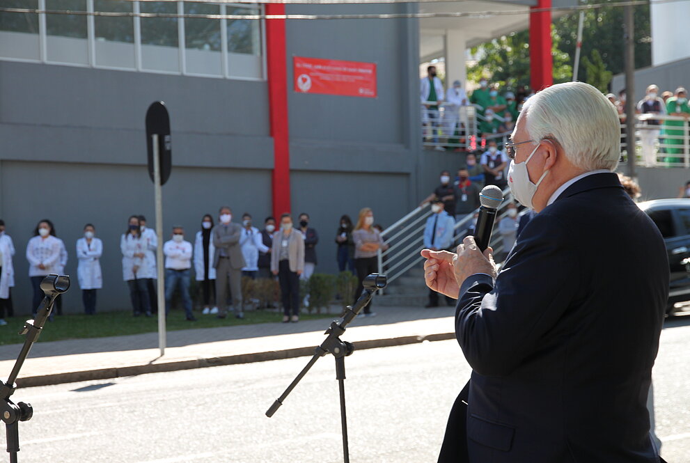 Homem de pé, usando máscara e segurando microfone discursa para várias pessoas ao fundo em frente à rua