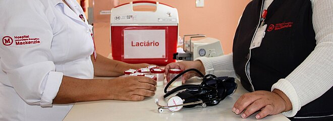 Na foto, as mãos de duas mulheres aparecem sobre um balcão branco onde estão apoiados um estetoscópio e uma caixa térmica vermelha