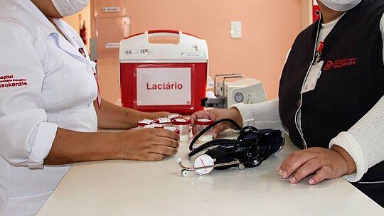 Na foto, as mãos de duas mulheres aparecem sobre um balcão branco onde estão apoiados um estetoscópio e uma caixa térmica vermelha