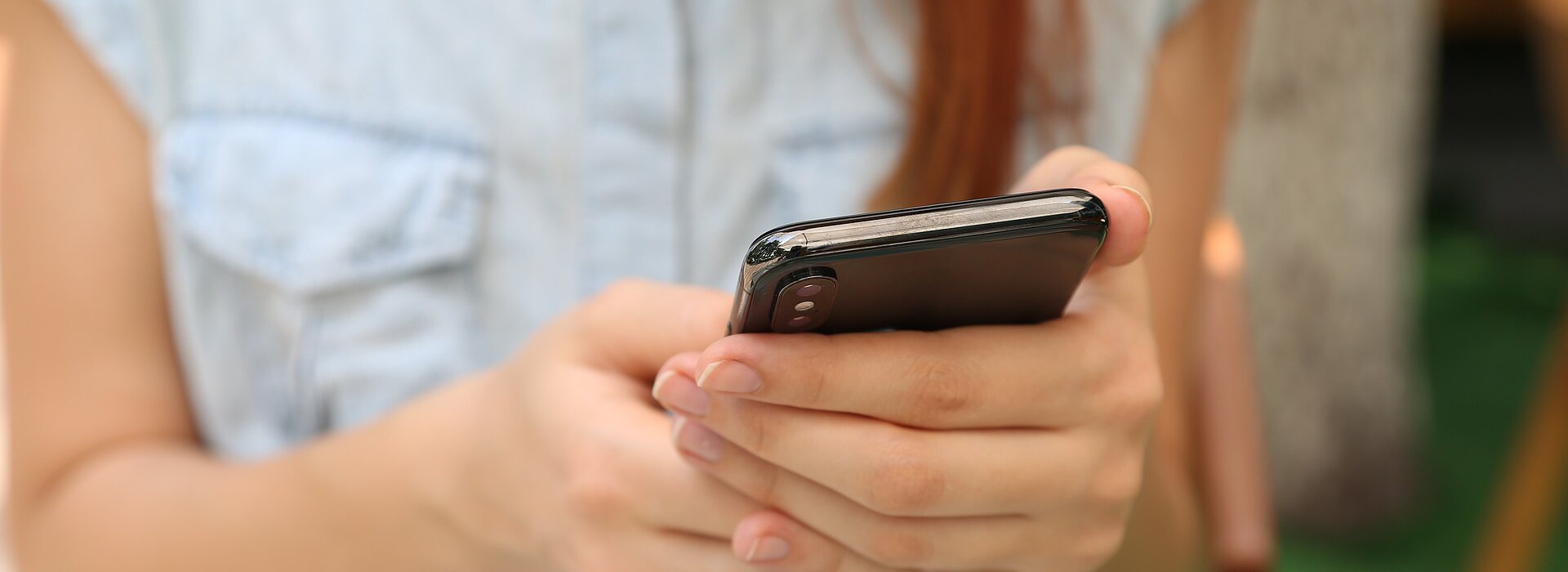 A foto mostra uma mulher com cabelo comprido mexendo no celular. Seu rosto não aparece, apenas seus braços e as mãos segurando o celular. 