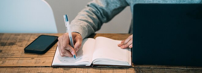 A foto mostra um homem, mas não conseguimos ver seu rosto. Ele está escrevendo em um caderno. Está com um celular do lado e um notebook na sua frente. 