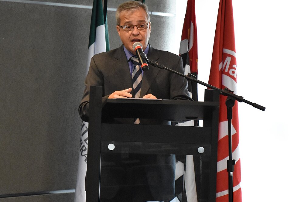 Pró-reitor está falando no pulpito em cima do palco do auditório do MackGraphe. Atrás estão algumas bandeiras.  