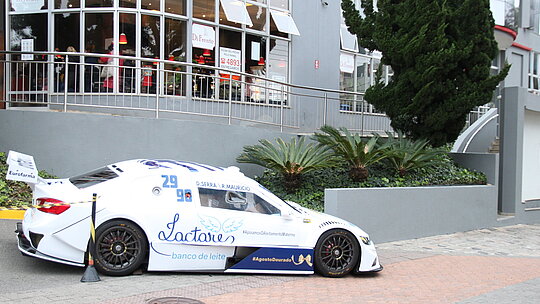 Carro branco da Stock Car estacionado em frente ao HUEM. Atrás pode-se ver o prédio cinza com logotipos em vermelho